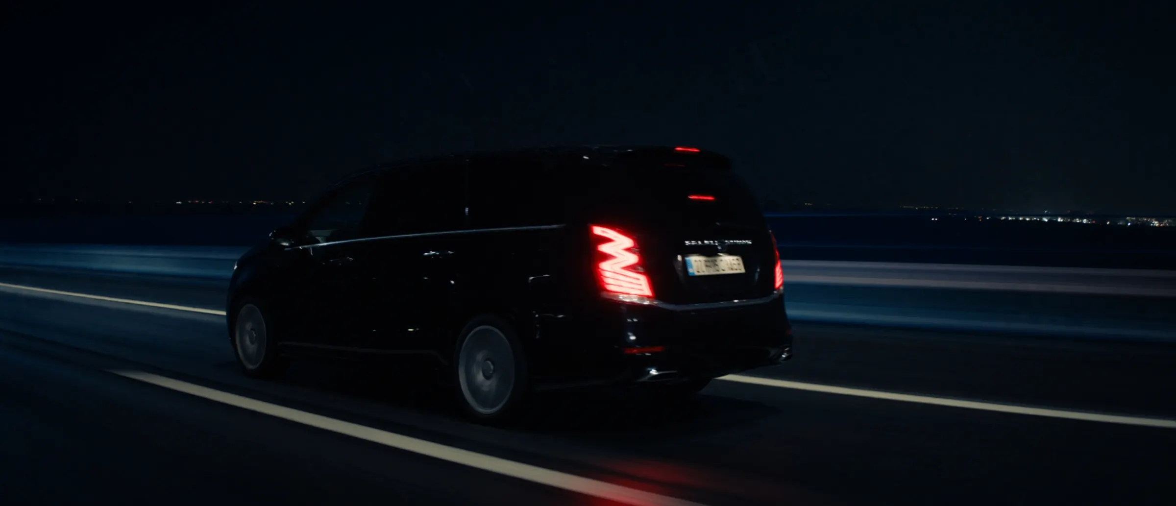 A black tour van on an empty motorway at night, tail lights streaking on wet asphalt
